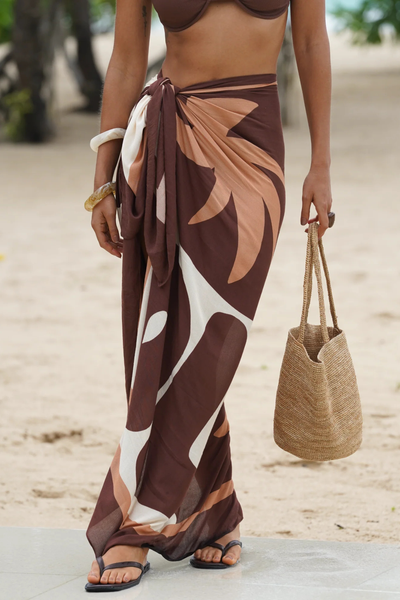 Mid-section detail shot of the Ochre Lane Sombra Sarong tied in a secure knot at the hip, highlighting the natural, fluid drape of the lightweight fabric. The close-up showcases the placement of the tan, cream, and chocolate brown abstract leaf print against the beach sand, accessorized with a woven straw bucket bag.