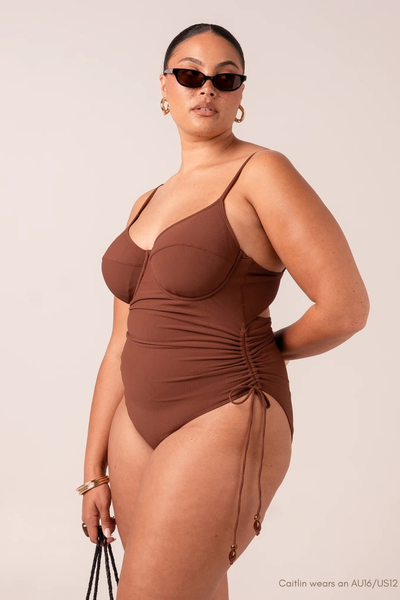 Studio front full-length view of plus-size model Caitlin wearing sunglasses and the chocolate brown underwire one-piece with adjustable side ties.