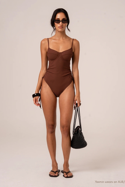 Studio front full-length view of model Yasmin wearing the chocolate brown underwire one-piece swimsuit, holding a black woven bag.