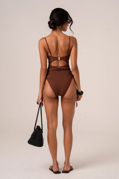 Studio rear full-length view of model Yasmin showcasing the open-back design, gold hardware clasp, and adjustable straps of the chocolate brown one-piece.