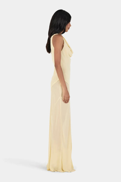 Studio side profile view of a model with long dark hair wearing a butter yellow draped cowl neck maxi gown featuring a panelled bias skirt.