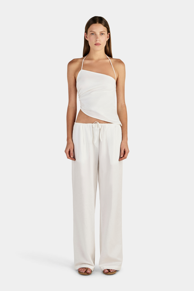 Studio full-length front view of a model wearing an ivory asymmetrical halter top with side gathering and an angled hem, paired with matching ivory wide-leg drawstring pants.
