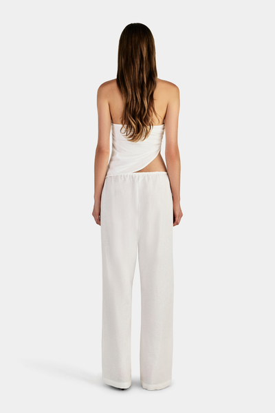 Studio full-length back view of a model showcasing the angled asymmetrical waist cut-out and low back design of an ivory halter top paired with white drawstring pants.