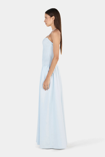 Studio side profile view of a model wearing a cornflower blue halter neck maxi dress with a fitted shirred bodice and a flowing full-length skirt.