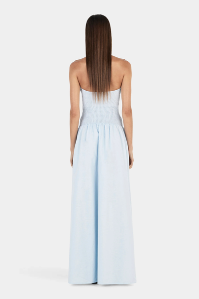 Studio rear full-length view of a model wearing a light cornflower blue maxi dress showcasing the smocked shirred back bodice and low cut.