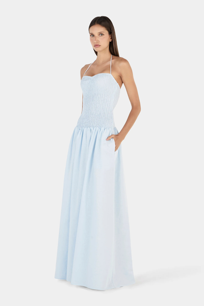 Studio three-quarter front view of a model with her hand in her pocket, wearing a cornflower blue halter maxi dress featuring a shirred bodice and gathered floor-length skirt.