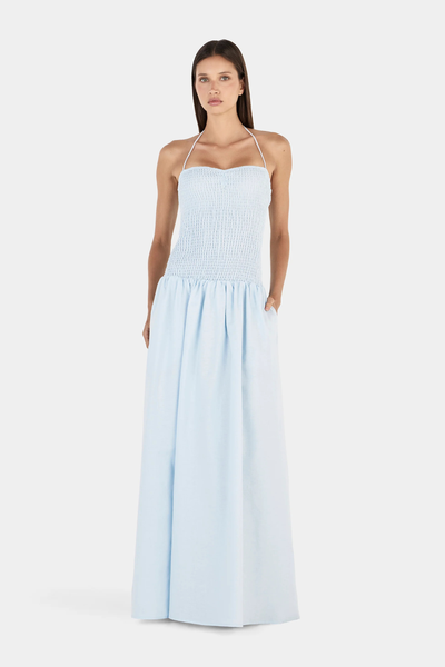 Studio full-length front view of a model wearing a light cornflower blue halter neck maxi dress with a fitted shirred bodice and a voluminous gathered skirt.