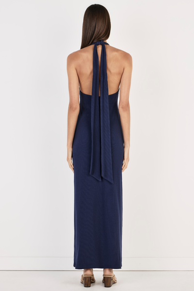 Back view of model wearing the Ownley Sachi Maxi Dress in Indigo, showcasing the open back design, long tied halter neck straps hanging down, and a fitted full-length ribbed jersey silhouette.