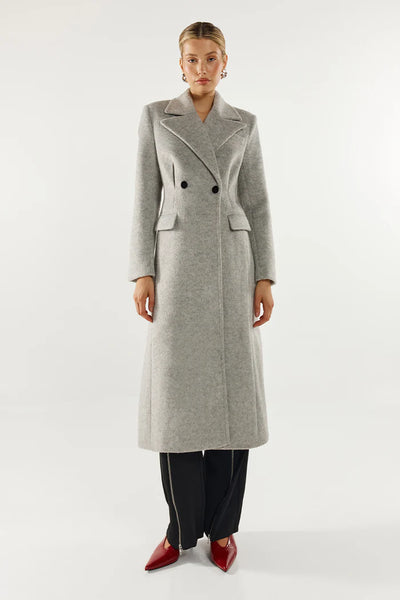 A rear three-quarter studio profile shot of a model wearing the grey longline Oliver Coat, emphasizing its structured shoulder pads, back vent detail, and the premium textured finish of the wool blend fabrication styled with wide-leg black trousers and red pointed-toe heels.