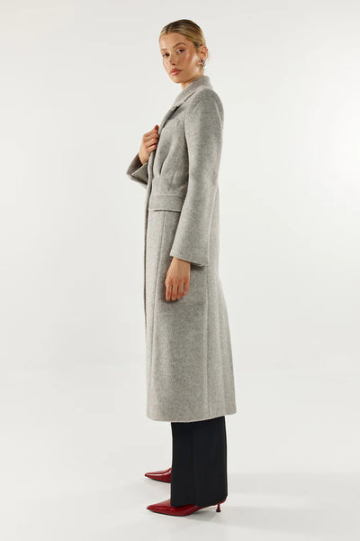 A full-length side profile studio image of the model highlighting the sharp tailoring, classic longline trench coat silhouette, and structured front flap pocket design of the grey winter coat.