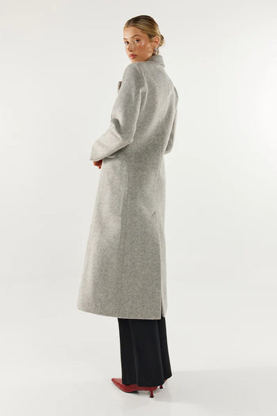 A full-length front studio shot of the model presenting the grey wool blend trench coat fully closed, showcasing its double-breasted design with two visible black buttons, integrated waist pleats, and an exaggerated notched collar and lapel.