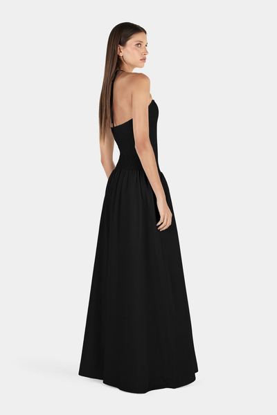 Studio rear three-quarter view of a model wearing a black halter-neck maxi dress with a fitted smocked bodice, thin neck straps, and a low open-back design.