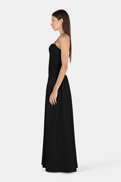 Studio side profile view of a model showcasing the form-fitting smocked bodice, delicate halter strap, and full-length gathered skirt of a black maxi dress.