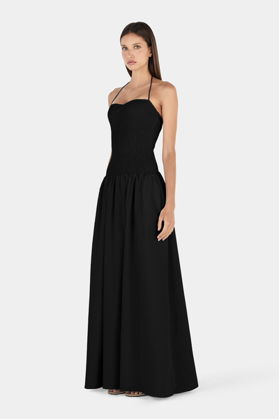 Studio three-quarter front view of a model posing with one hand in her pocket, wearing a black smocked halter-neck maxi dress.