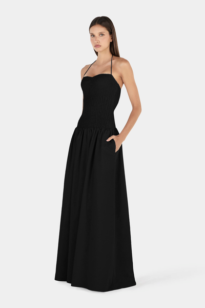 Full-length studio front view of a model with her left hand in her pocket, wearing a black halter-neck maxi dress featuring a shirred bodice and full gathered skirt.