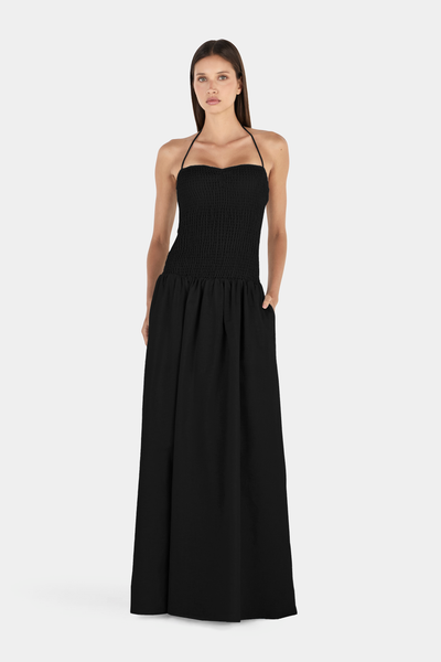 Studio full-length front view of a model posing with both hands in her side pockets, wearing a black smocked halter maxi dress.