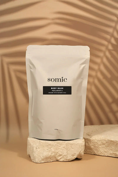 A white stand-up pouch of somie body wash with a black label, displayed on a textured stone block against a warm beige background with leaf shadows.
