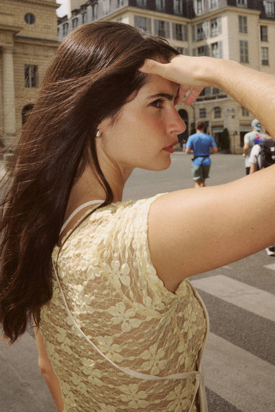 Close-up side profile portrait of a model with dark hair wearing the creme wildflower lace cap-sleeve top against an outdoor city backdrop.