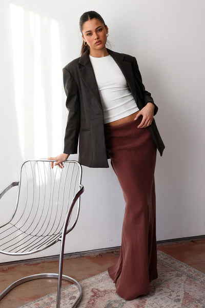 An indoor lifestyle campaign image of a model leaning against a white studio wall beside a metallic wire chair, wearing the chocolate brown PU leather blazer over a white fitted baby tee and paired with a flowing chocolate brown maxi skirt.