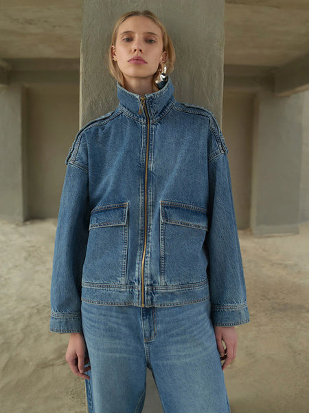 A close-up campaign shot of a model wearing the Kivari Rita Denim Jacket fully zipped, showcasing the high structured funnel neck collar design and oversized patch pockets.