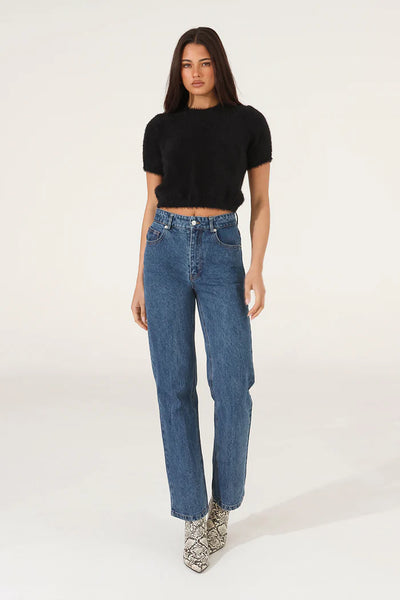 A full-length front studio shot of a model wearing the black fluffy knit tee, showcasing its cropped hemline, short sleeves, and relaxed scooped neckline styled with high-waisted blue jeans and snakeskin boots.