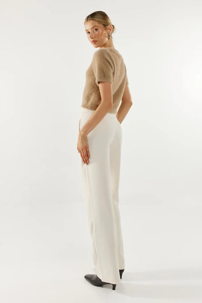 A full-length studio profile shot from the rear angle, showcasing the clean back drape and short-sleeve construction of the Ruby fluffy knit tee, styled with tailored high-waisted cream trousers and black pointed-toe heels.