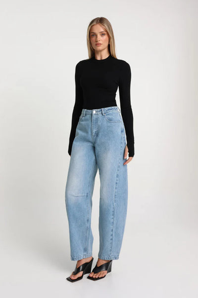 A full-length rear studio view of a model wearing the black fitted long-sleeve top paired with high-waisted, light-wash balloon jeans and black kitten heel mules.