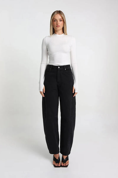 A full-length studio shot of the model standing forward-facing in the white fitted long-sleeve top, showcasing its seamless stretch fabrication and thumbhole sleeve details paired with wide-leg black jeans and black open-toe wedge heels.
