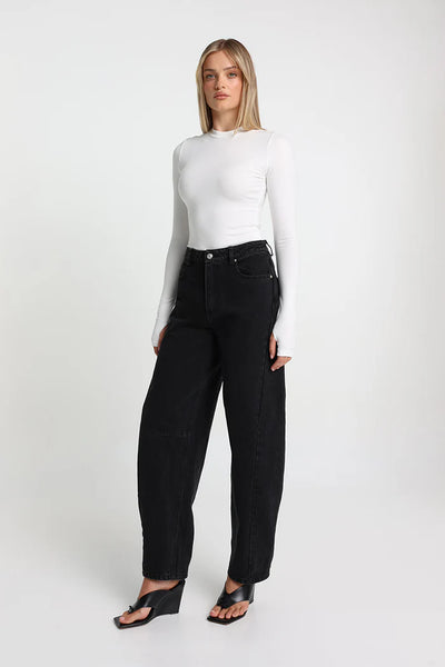A full-length rear profile studio view highlighting the clean, figure-hugging fit of the white long-sleeve baby tee styled with high-waisted black balloon denim.