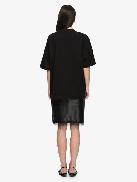 Full back view studio shot of a model wearing the Bec + Bridge Saphira Midi Skirt in Black faux leather with delicate lace hem trim, styled with an oversized black t-shirt and heels.