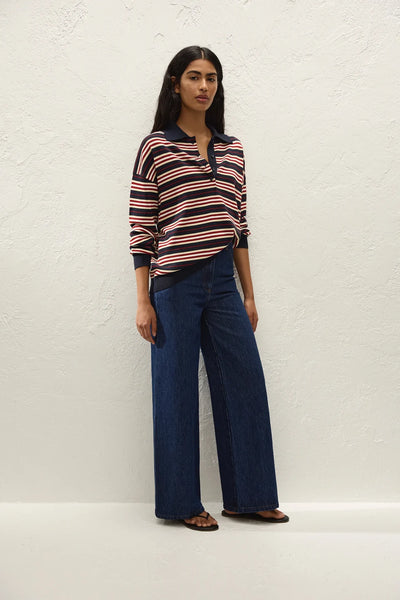 Full-length front view of model posing in the long-sleeve Faithfull The Brand Harlow Oversized Polo in Cila Stripe Navy, dark blue wide-leg jeans, and black sandals.
