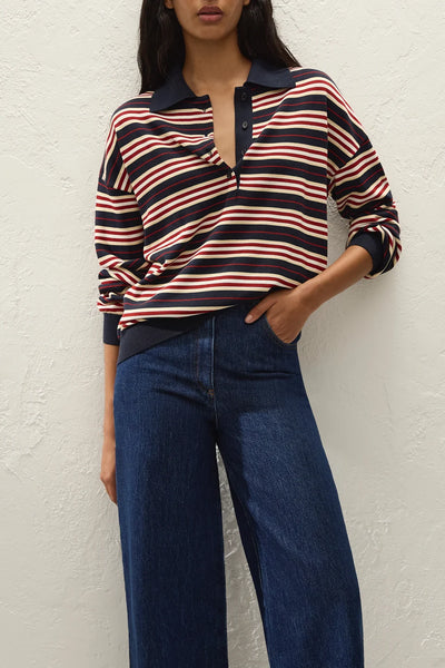 Three-quarter torso view of model wearing the Faithfull The Brand Harlow Oversized Polo in Cila Stripe Navy with open button placket and hand in jean pocket against a white wall background.