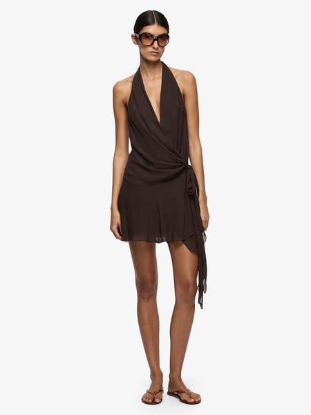 A full-length front studio photograph of the model standing against an isolated white background. This framing captures the complete silhouette of the dark chocolate 100% viscose dress, detailing the wrap-style bodice, the asymmetrical draped wrap skirt line with soft gathers, and the lightweight sheer polyester-lined hemline, paired with flat brown thong sandals.