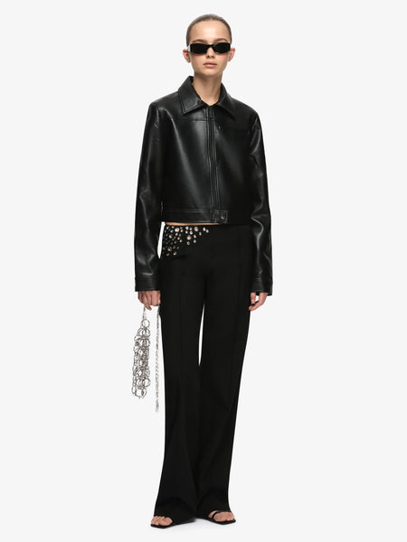 Full-length front view studio portrait of a model wearing the Bec + Bridge Saphira Cropped Jacket in Black faux leather, paired with black flared pants, sunglasses, and a silver chain bag.