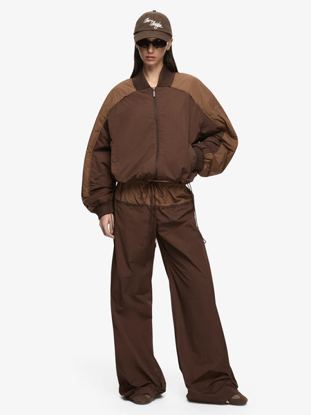 Full-length front view studio portrait of a model wearing the Bec + Bridge Laney Jacket in Choc/Coffee styled with matching two-tone track pants, a brown cap, and sunglasses.