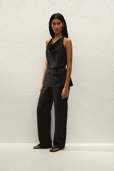Full-length front view of a model wearing a black cowl-neck halter top tucked into the matching fluid trousers, paired with thong sandals.