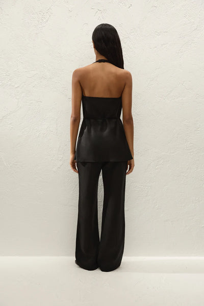 Full-length rear view of a model wearing a matching black halter top and the Varina trousers with sandals against a textured white wall.