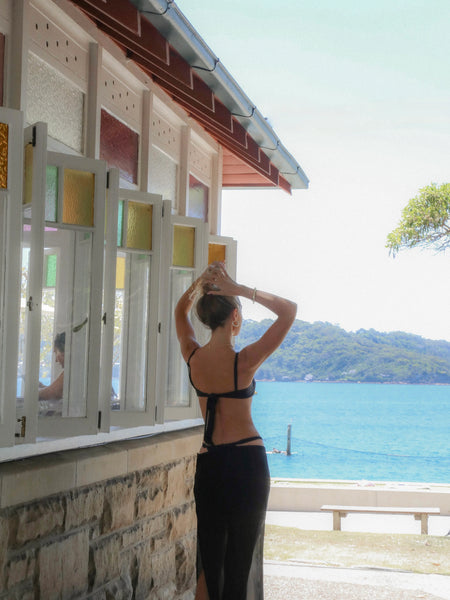 View of a model with her hands clasped behind her head, standing near a building with stained-glass windows overlooking the blue sea.
