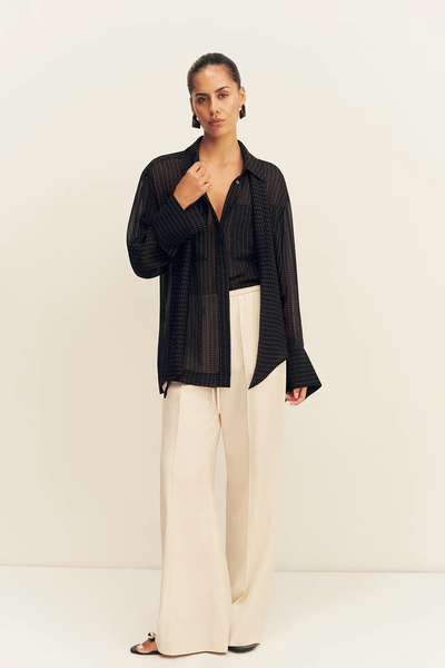 Front studio view of a model posing in the Shona Joy Irving Polka Dot Sheer Shirt in Black Polka Stripe, styled open over cream pants.