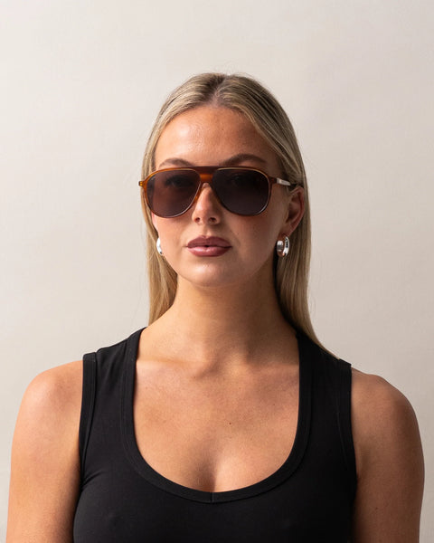 Front portrait of a blonde model wearing the Sito Shades Bobbi in Tangelo translucent orange aviator sunglasses paired with silver hoop earrings and a black tank top.