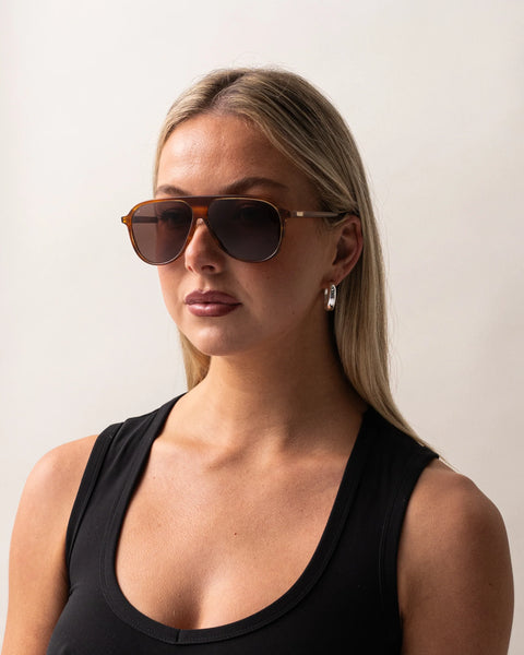 Front portrait of a blonde model wearing the Sito Shades Bobbi in Tangelo translucent orange aviator sunglasses paired with silver hoop earrings and a black tank top.