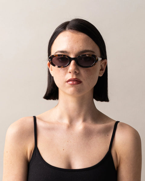 Front portrait of a dark-haired model wearing the Sito Shades Lourdes oval sunglasses in pebble tort paired with a black camisole top against a plain studio background.