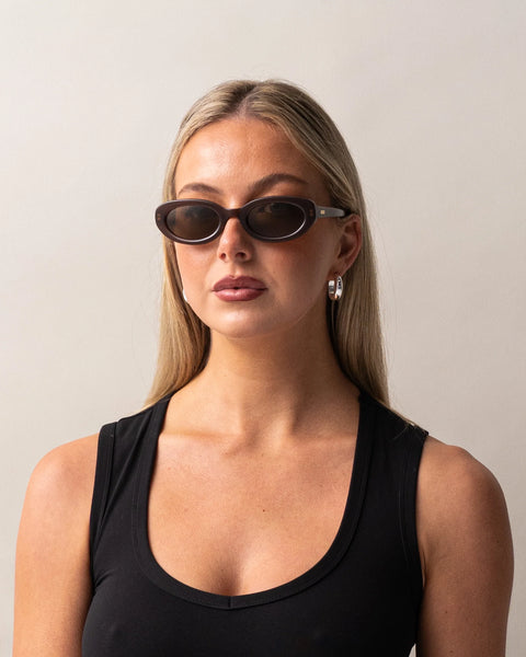 Front portrait of a blonde model wearing the Sito Shades Lourdes oval sunglasses in milky coco brown styled with a black tank top and silver hoop earrings.