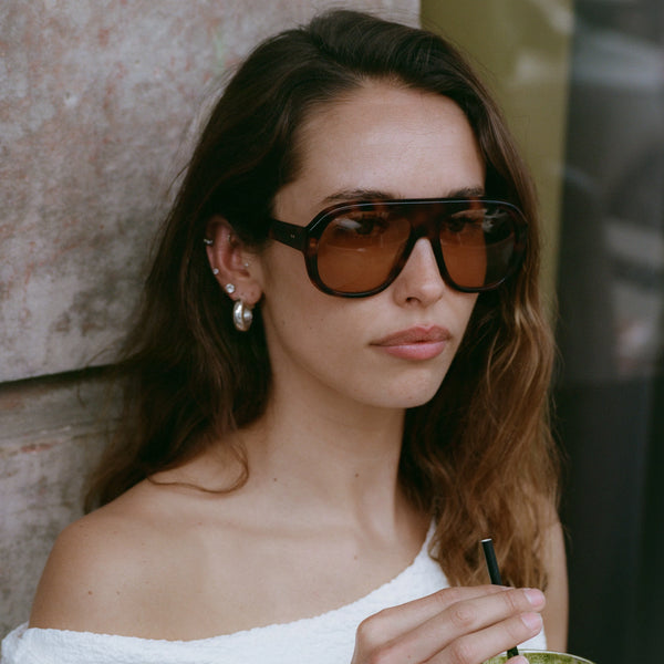 Outdoor lifestyle portrait of a woman with long dark wavy hair wearing oversized translucent classic tortoiseshell aviator-style sunglasses with amber-tinted lenses, an off-the-shoulder top, and hoop earrings.