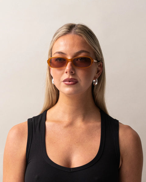 Front portrait of a blonde model wearing the Sito Shades Roux rectangular sunglasses in translucent caramel orange styled with a black tank top and silver hoop earrings.