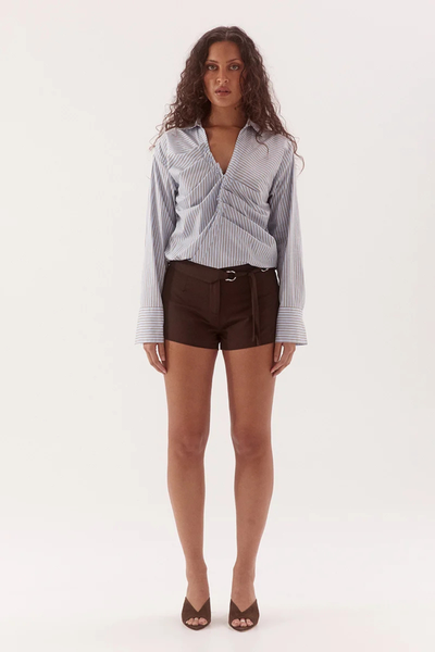 Full-length studio front view of a model wearing a long-sleeve light blue and brown pinstripe fixed-wrap cotton shirt featuring a V-neckline, tucked placket detailing, and wide cuffs, styled with dark brown high-waisted shorts.