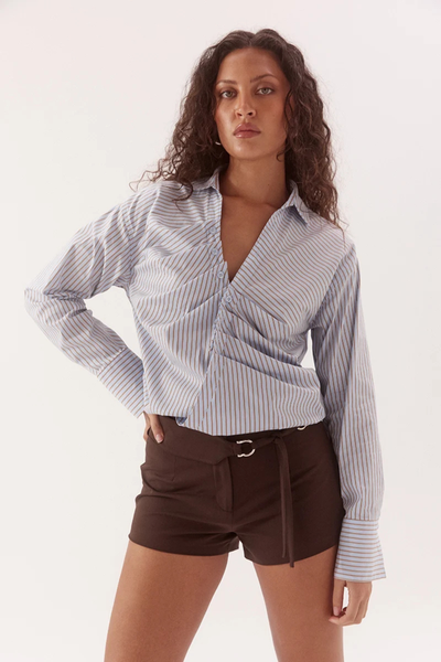Studio three-quarter front portrait of a model wearing a blue and brown striped long-sleeve wrap shirt with front placket tuck details, wide sleeve cuffs, and dropped shoulders paired with brown shorts.