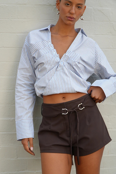 Close-up outdoor shot of a model against a light wall wearing a blue and brown pinstripe wrap shirt highlighting the front neckline tuck details, open V-neck placket, and wide cuffs.