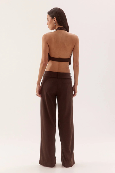 Full-length studio rear view of a model wearing an open-back dark chocolate apron top with a button-fastened halter neck and horizontal back strap, paired with matching brown tailored pants.