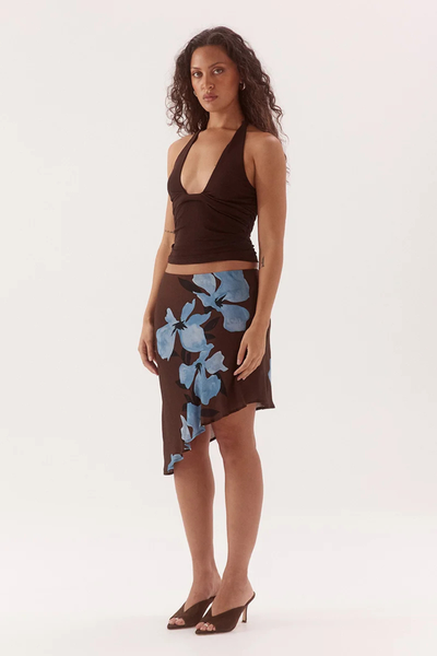 Studio three-quarter front view of a model wearing a dark chocolate brown stretch mesh halter top with a front scoop neckline and waist gathers, styled with an asymmetrical floral print brown skirt and dark heels.